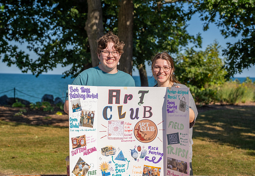 Two students in front of a board that is promoting the campus Art Club.
