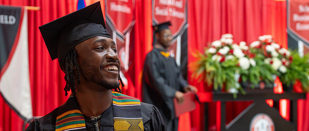 A student in their graduation robes smiling in front a the stage.