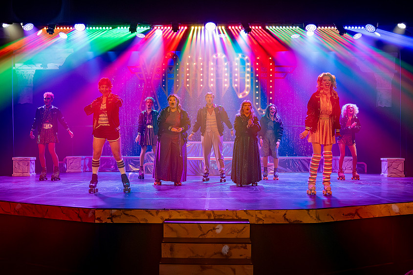 Multiple student actors on the stage set as a roller disco. Different colored spotlights highlight the stage.