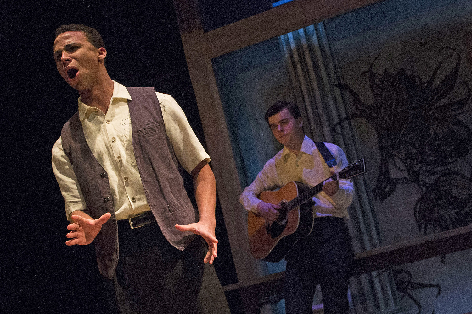 A person singing while a person in the background plays guitar.