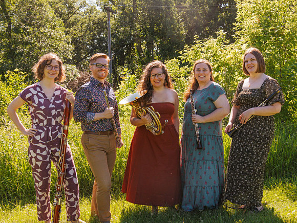 A group of musicians holding their instruments in a wooded outdoors area.